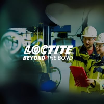 Kaksi tehdastyöntekijää suojavaatteissa ja kypärät päässä. He tarkastelevat konelaitteistoa ja punaista tablettia teollisessa ympäristössä. Kuvan keskellä näkyy LOCTITE-logo ja iskulause "BEYOND THE BOND"