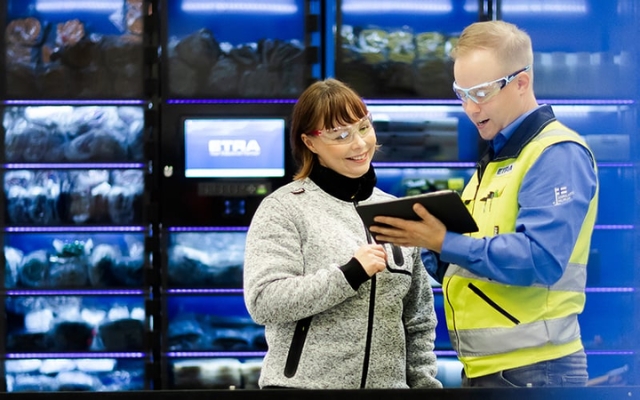 Etra specialist presenting products to a customer on a tablet in front of an automated storage system, both wearing safety glasses.