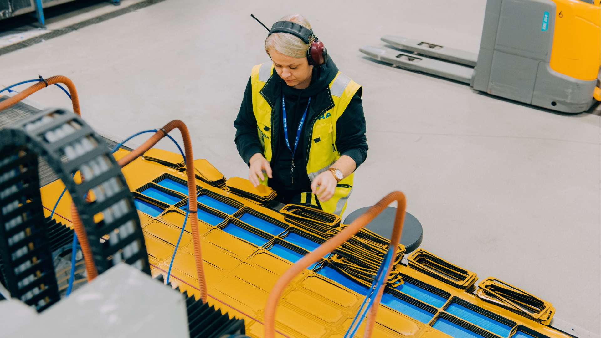 A person wearing a yellow safety vest and headphones is working with rubber material. A forklift is visible in the background.
