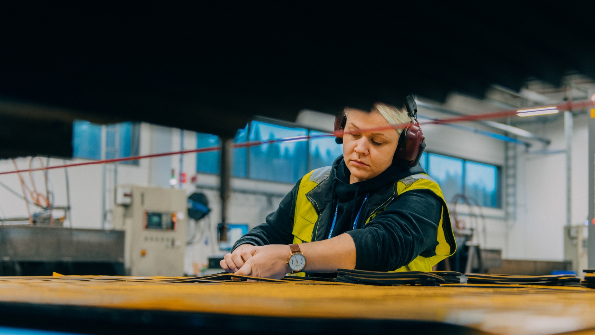 A person wearing a high-visibility vest and ear protection is working at a table in an industrial or workshop setting. Large windows and various equipment and machinery are visible in the background.