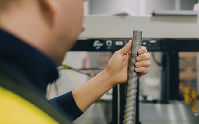 Worker holding a threaded metal rod in front of a production machine, close-up view.