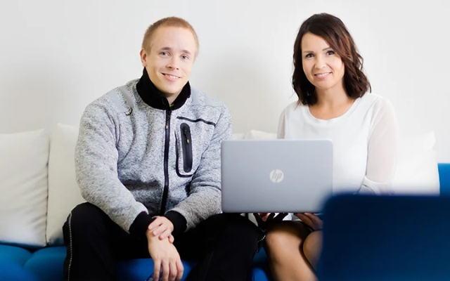 Two people sitting on a sofa with a laptop, smiling at the camera in a professional setting.