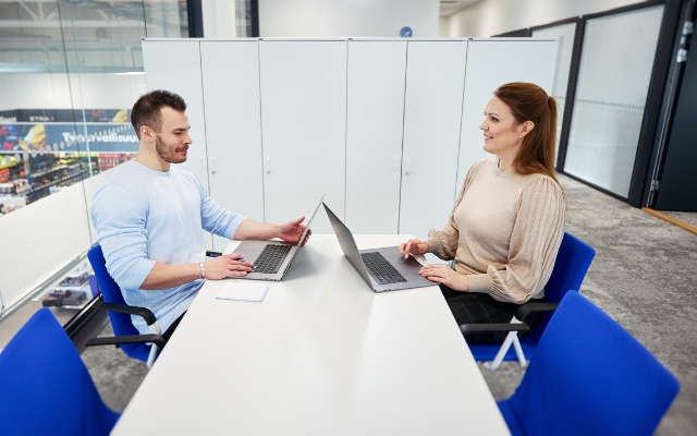 A man and a woman are sitting across from each other in an office at a conference table, each with a laptop in front of them. They are smiling while having a conversation.