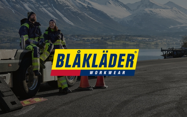 Kaksi henkilöä yllään Blåkläderin työvaatteet istuu perävaunun reunalla talvisessa vuoristomaisemassa järven rannalla. Kuvan keskellä on Blåkläder Workwear -logo keltaisella, sinisellä ja punaisella taustalla.