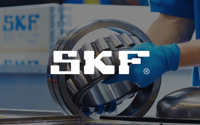 Henkilö sinisissä suojakäsineissä käsittelee suurta laakeria työpisteellä. Taustalla näkyy SKF-logoja laatikoissa. Kuvan keskellä on suuri valkoinen SKF-logo.
