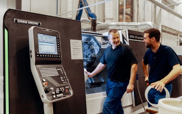 Två arbetare står vid en CNC-maskin i en plastproduktionsanläggning och samtalar. I förgrunden finns en DMG MORI-kontrollpanel och i bakgrunden en CTX 510 ecoline-svarv. Arbetarna har mörkblå arbetskläder på sig.