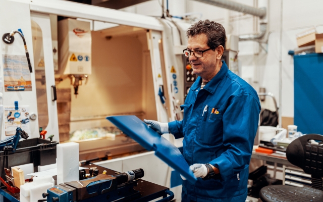 En arbetare i blå arbetskläder hanterar en blå plastskiva framför en CNC-maskin i en plastproduktionsanläggning. Arbetsstationen har verktyg, plastdelar och en verktygslåda.