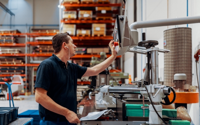 En arbetare i en plastproduktionsanläggning startar eller stoppar en maskin genom att trycka på den röda nödstoppknappen på kontrollpanelen. I bakgrunden finns lagringshyllor, plastdelar och bearbetningsutrustning.