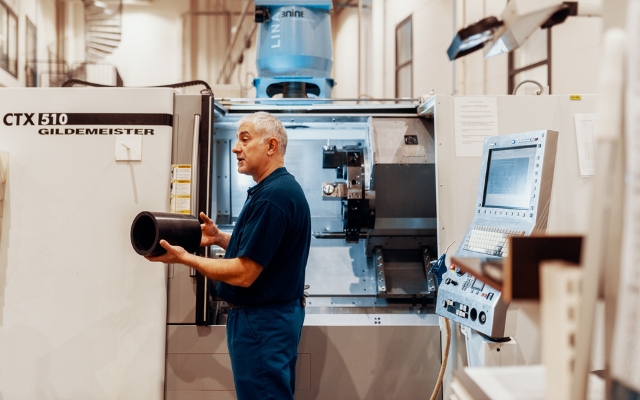 En arbetare i en plastproduktionsanläggning håller ett svart plaströr framför en CNC-maskin. På maskinens sida står det "CTX 510 GILDEMEISTER", och bredvid finns en kontrollpanel.