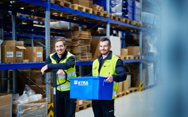 Varastotyöntekijät hymyilevät ja työskentelevät logistiikkakeskuksessa, pukeutuneina keltaisiin huomioliiveihin. Toinen työntekijä pitelee sinistä muovilaatikkoa, jossa lukee 'ETRA – Your Industrial Partner', kun taas toinen nojaa pumppukärryyn.