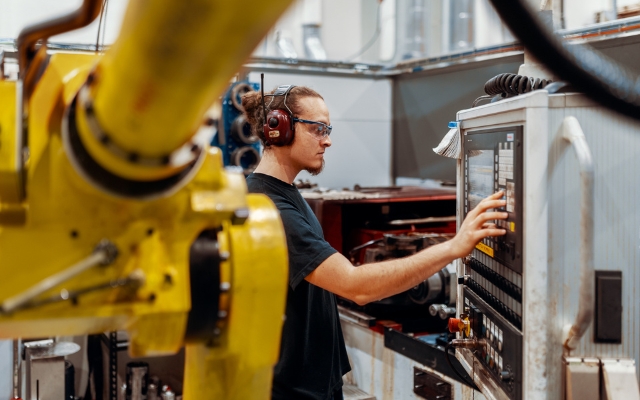 A worker operates the control panel of a CNC machine in an industrial facility. The person is wearing safety glasses and hearing protection. In the foreground, a yellow robotic arm is visible.