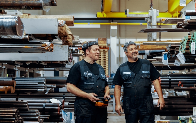 Two workers stand in a metal storage area between shelves filled with metal bars and pipes of varying lengths and thicknesses. One worker is holding a remote control, and both are wearing black work clothes and vests. In the background, a yellow crane is 