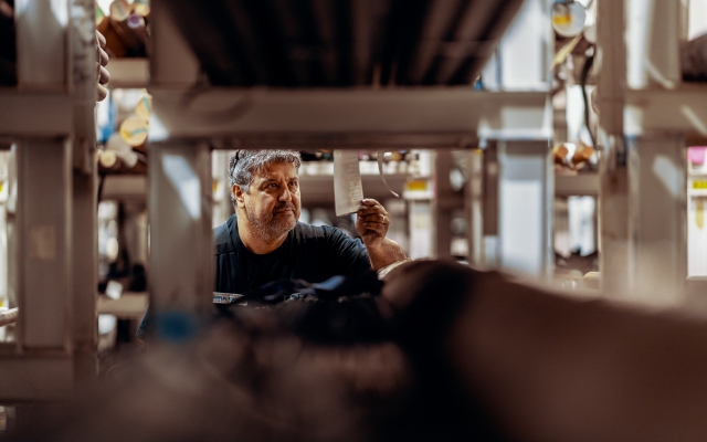 A worker in a metal storage area examines a label attached to a metal pipe between the shelves. The shelves contain metal bars and pipes of various sizes. In the background, rows of storage shelves are visible.