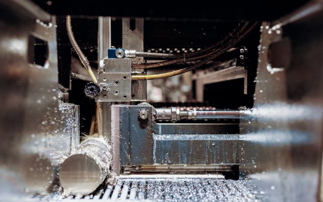 En metallisk sågmaskin skär en rund metallstång i en produktionsanläggning. Skärprocessen producerar metallspån och kylvätska strömmar runt sågbladet. Röret är fastsatt i en robust hållare inne i maskinen.