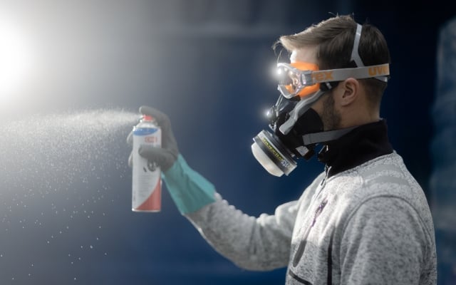En arbetare med skyddsmask, skyddsglasögon och handskar sprayar smörjmedel från en aerosolförpackning. Dimman belyses av starkt ljus mot en mörk bakgrund.