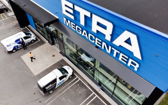 Flygvy av ETRA Megacenter-byggnaden med en stor blå fasad och tydlig vit text. Leveransskåpbilar är parkerade framför, och en person i orange väst går på trottoaren. Byggnaden har stora glasfönster som visar interiören.