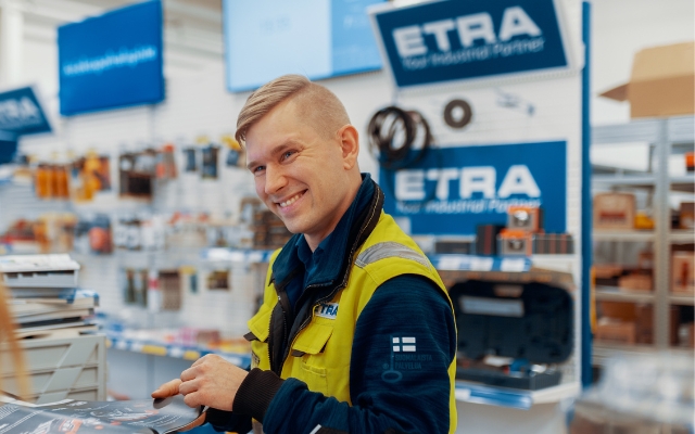 Hymyilevä mies asiakaspalvelija seisoo myymälässä ETRA-kyltin edessä, päällään keltainen huomioliivi ja sininen työasu, taustalla hyllyjä täynnä työkaluja ja tarvikkeita.