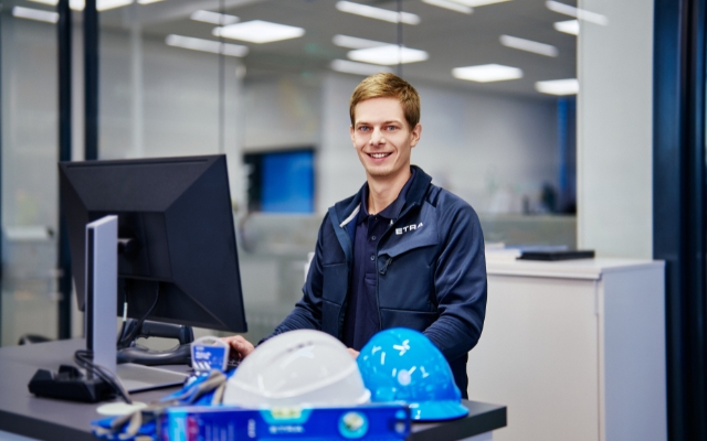 An office worker smiles at his workstation in a modern office environment. He is wearing a dark blue ETRA work jacket and working on a computer. On the desk, there is a blue and white safety helmet, a toolbox, and a blue coffee mug with the ETRA logo.