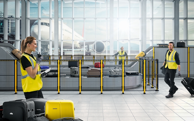Airport baggage handling area where staff in safety vests are sorting and handling luggage, with a passenger airplane visible in the background.