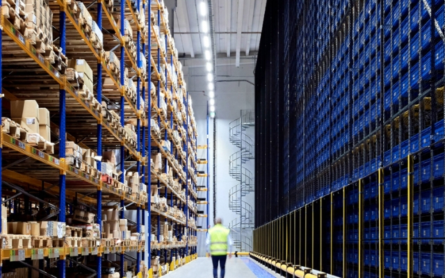 Moderni varastohalli, jossa korkeat hyllystöt, sinisiä laatikoita automaatiojärjestelmässä ja henkilö huomioliivissä kävelemässä käytävällä.