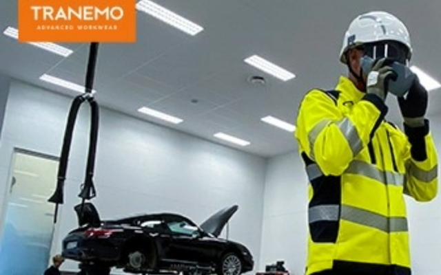 Korjaamotyöntekijä heijastavassa työvaatteessa ja suojakypärässä, autohuolto käynnissä taustalla"