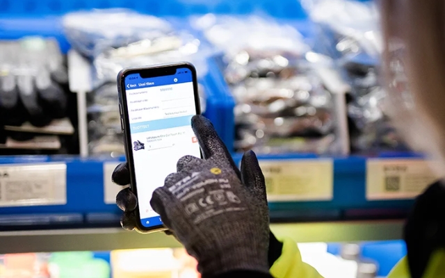En person med handskar beställer produkter med en smartphone i en lagermiljö, med hyllor och skyddsutrustning i bakgrunden.