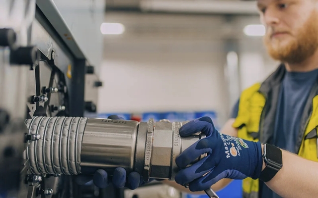 Worker installing an industrial hose fitting to a machine, wearing protective gloves and a high-visibility vest.