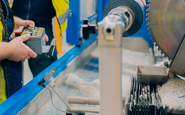 Worker operating an industrial saw machine using a control panel, wood shavings and spinning blade visible in the work area.