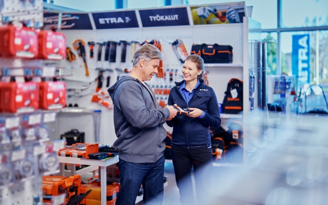 Asiakaspalvelija auttaa asiakasta työkaluliikkeessä. Nainen, jolla on ETRA-työvaatteet, hymyilee ja esittelee tuotetta harmaapaitaiselle miehelle. Taustalla on hyllyjä, joissa on erilaisia työkaluja ja tarvikkeita.