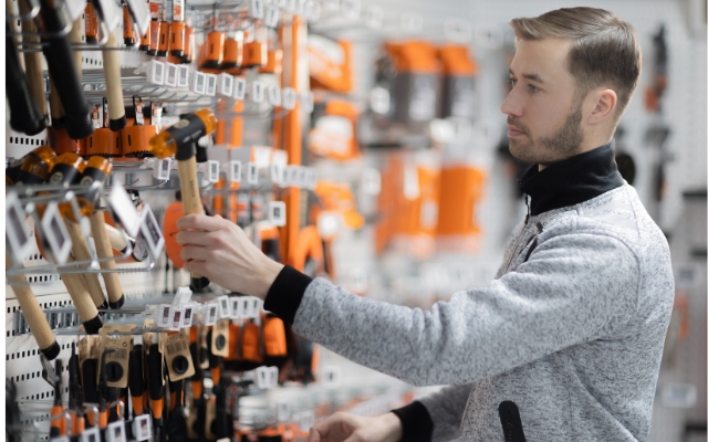 Mies tarkastelee vasaraa työkaluliikkeen hyllystä. Hänellä on harmaa takki ja hän seisoo hyvin valaistussa myymälässä, jossa on erilaisia työkaluja oransseilla ja mustilla pakkauksilla.