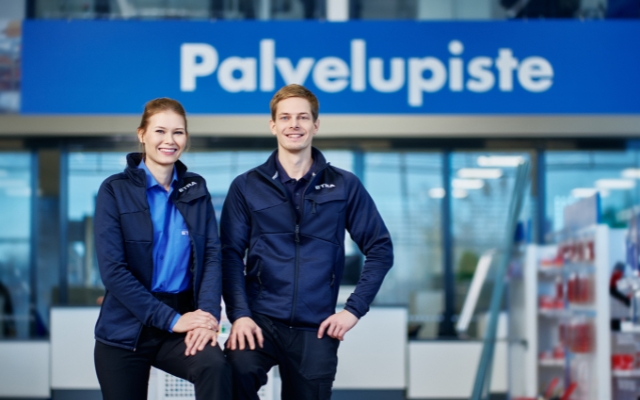 Kaksi hymyilevää asiakaspalvelijaa seisoo palvelupisteen edessä. Heillä on päällään siniset työasut, ja taustalla näkyy moderni myymäläympäristö kirkkaassa valaistuksessa.
