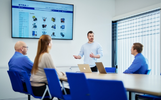 A presentation in a meeting room where a young man is standing and speaking to three seated individuals. In the background, a large screen displays a website showcasing various tools.