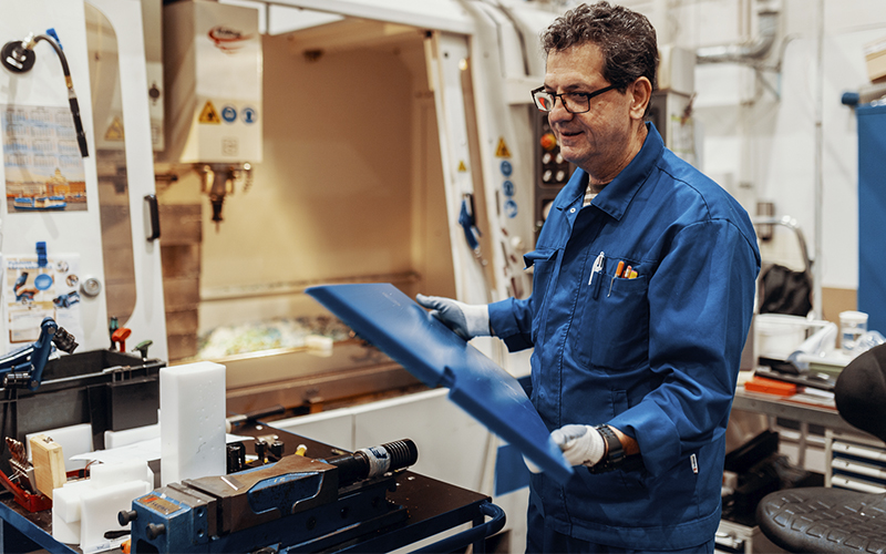 En arbetare i blå arbetskläder hanterar en blå plastskiva framför en CNC-maskin i en plastproduktionsanläggning. Arbetsstationen har verktyg, plastdelar och en verktygslåda.