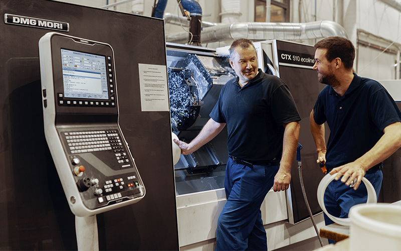 Två arbetare står vid en CNC-maskin i en plastproduktionsanläggning och samtalar. I förgrunden finns en DMG MORI-kontrollpanel och i bakgrunden en CTX 510 ecoline-svarv. Arbetarna har mörkblå arbetskläder på sig.