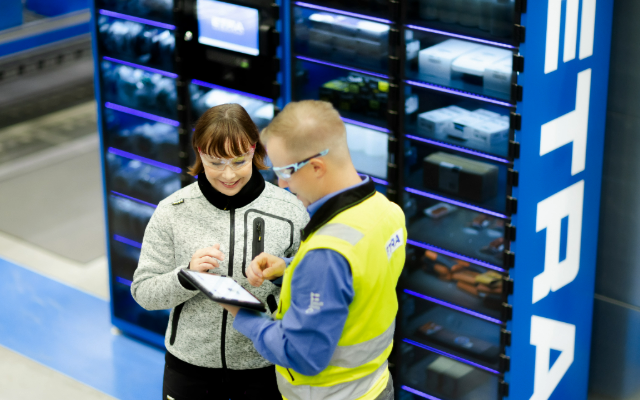 Two people stand in front of an Etra eLock service storage cabinet. They are wearing safety glasses, and one person, dressed in a high-visibility vest, is using a tablet while discussing with the other.