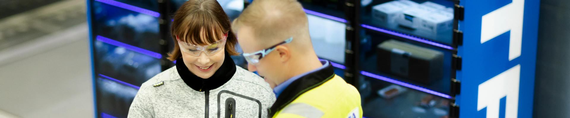 Två personer står framför ett Etra eLock-servicelager. De bär skyddsglasögon, och en av dem, klädd i en varselväst, använder en surfplatta medan de diskuterar med den andra personen.
