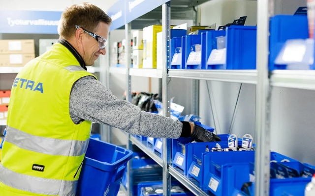 An Etra employee picks products from blue storage bins on a shelf in a warehouse environment, wearing a high-visibility vest and safety glasses.