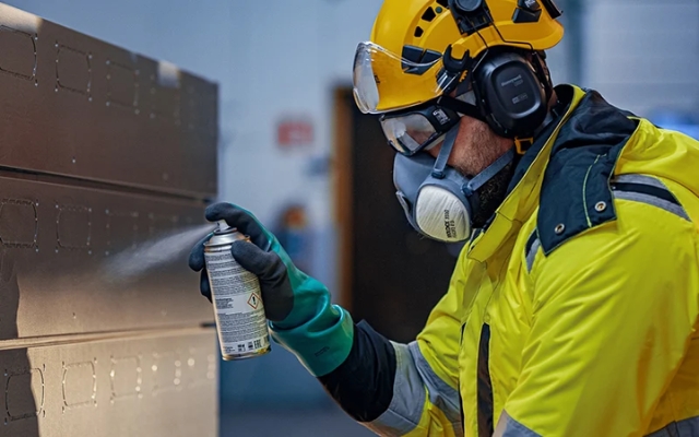 Worker in protective gear spraying aerosol onto a metal surface, wearing a respirator, hearing protection, and safety gloves.