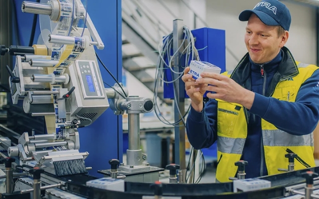 Etra-anställd som kontrollerar en produkt vid en förpackningsmaskin i en produktionsmiljö, iförd varselväst och keps.