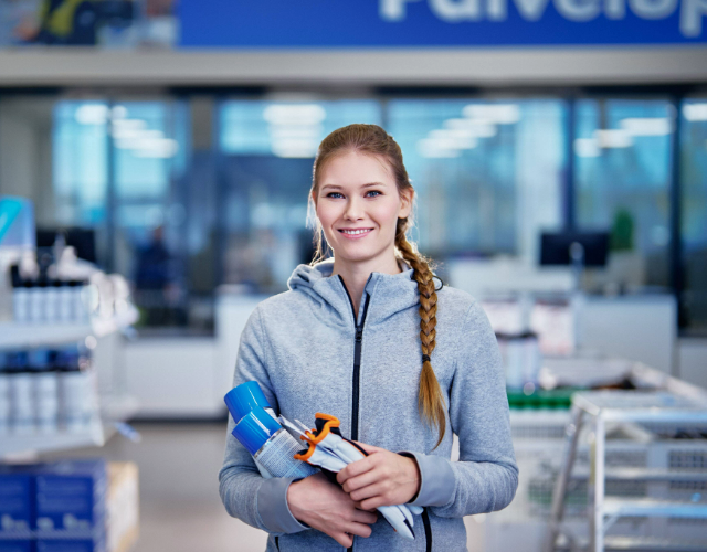 Hymyilevä nainen seisoo myymälässä pitäen käsissään erilaisia tuotteita, kuten suihkepulloa ja työkalupakkauksia. Hänellä on harmaa huppari, ja taustalla näkyy hyllyjä ja kassapisteitä kirkkaasti valaistussa tilassa.