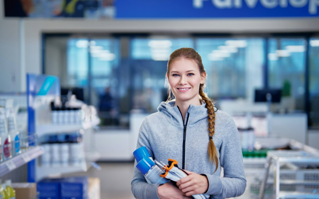 Hymyilevä nainen seisoo myymälässä pitäen käsissään erilaisia tuotteita, kuten suihkepulloa ja työkalupakkauksia. Hänellä on harmaa huppari, ja taustalla näkyy hyllyjä ja kassapisteitä kirkkaasti valaistussa tilassa.