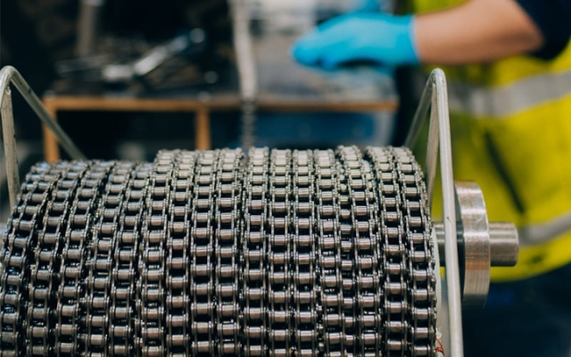 Close-up of an industrial chain reel in a production area, with a person wearing protective gloves and a high-visibility vest in the background.