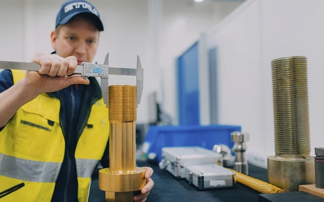 Etra employee measuring a large threaded metal part with a caliper at a workstation.