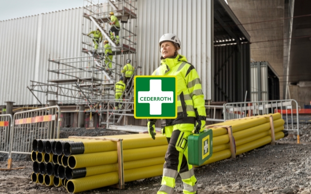 Rakennusty&ouml;maalla seisova ty&ouml;ntekij&auml; pukeutuneena keltaiseen heijastavaan suojavaatetukseen ja valkoiseen kyp&auml;r&auml;&auml;n. H&auml;n kantaa vihre&auml;&auml; ensiapulaukkua, jossa on Cederroth-logo. Taustalla n&auml;kyy rakennustelineit&auml;, muita ty&ouml;ntekij&ouml;it&auml; ja keltaisia suojaputkia
