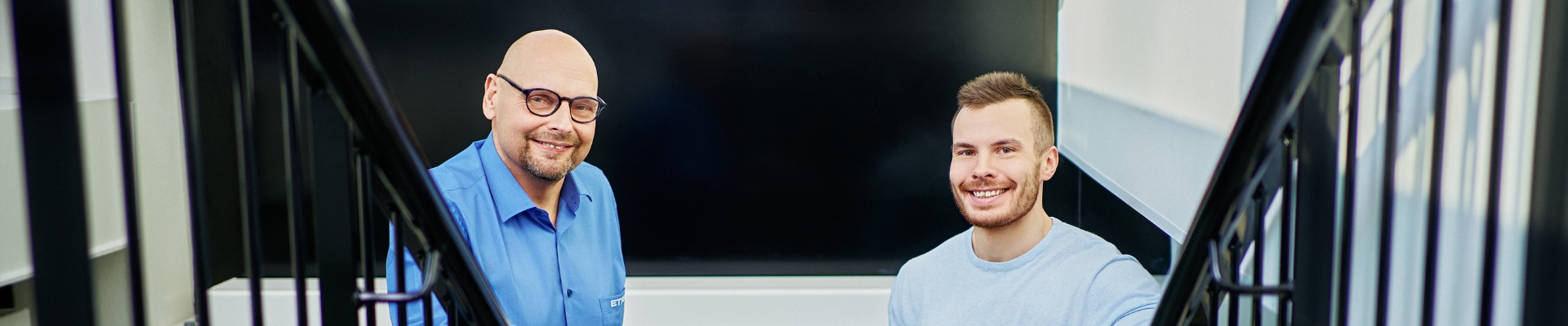 Two smiling men are standing outside on a staircase, leaning on the railing. One is wearing a blue dress shirt and glasses, while the other is wearing a light blue t-shirt.