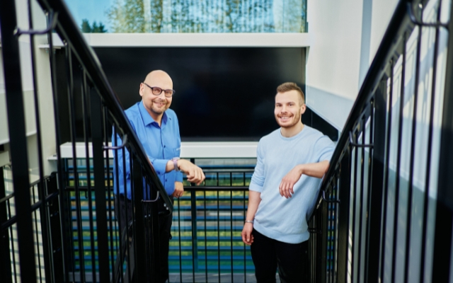 Two smiling men are standing outside on a staircase, leaning on the railing. One is wearing a blue dress shirt and glasses, while the other is wearing a light blue t-shirt.